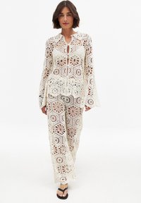 Abito in due pezzi all'uncinetto color bianco sporco con motivi floreali, maniche lunghe, colletto e pantaloni a gamba larga. Indossato con sandali neri.