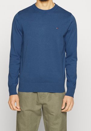 Uomo che indossa un maglione blu a maniche lunghe con scollo rotondo e un piccolo logo Tommy Hilfiger sul petto, abbinato a pantaloni verde oliva.