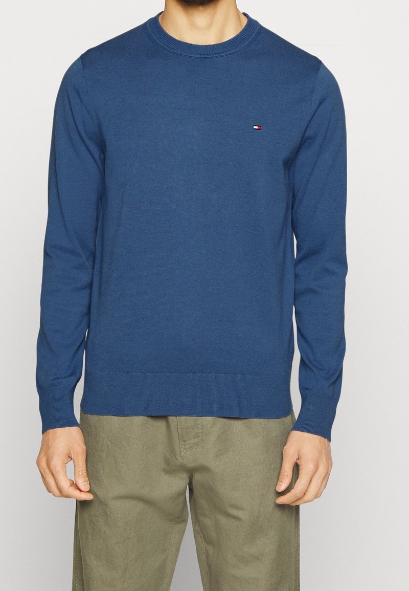 Homme portant un pull bleu à col rond à manches longues avec un petit logo Tommy Hilfiger sur la poitrine et un pantalon vert olive.