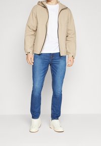 Beige kevytrakenteinen takki hupulla, vetoketjullinen etuosa, yhdessä valkoisen t-paidan ja sinisten farkkujen kanssa. Valkoiset lenkkarit täydentävät asua.