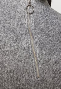 Grijs gebreide trui met een halve rits voorzien van een zilveren ringtrekker. De stof heeft een gestructureerd patroon en geeft een gezellige uitstraling.
