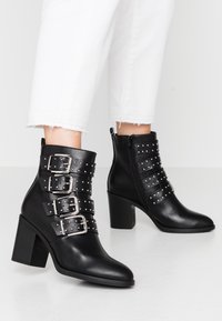 Botas de tobillo de cuero negro con cuatro correas con hebillas, acentos de remaches plateados, punta afilada y un tacón bloque grueso. Cierre de cremallera lateral.