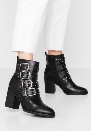 Black leather ankle boots with four buckled straps, silver rivet accents, pointed toe, and a chunky block heel. Side zipper closure.