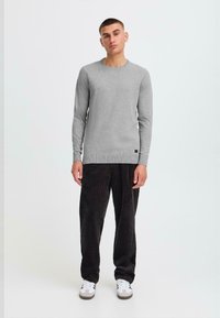 Young man standing, wearing a gray long-sleeve sweater, black corduroy pants, and white sneakers with black stripes, against a plain background.