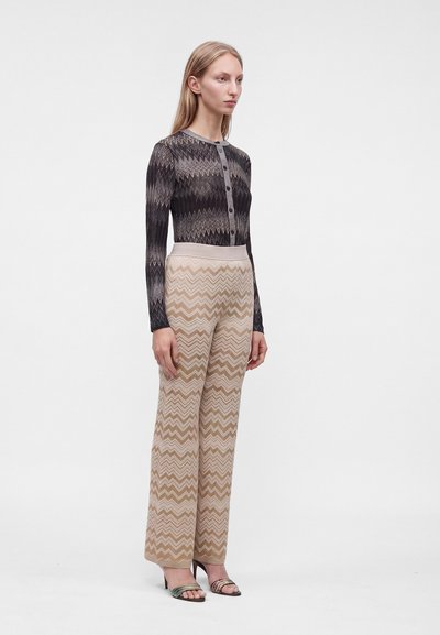 Femme debout portant un haut boutonné à motif noir et gris, un pantalon large beige à motif zigzag et des sandales à talons hauts.
