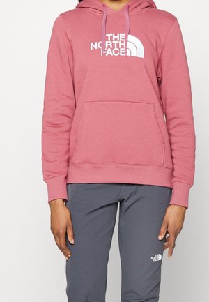 Person wearing a dusty pink hoodie and dark gray pants, both with white The North Face logo, standing against a plain background.