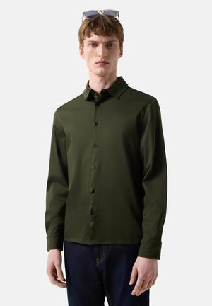 Junger Mann trägt ein dunkelolivgrünes Hemd mit Knöpfen und dunkle Jeans, mit Sonnenbrille auf dem Kopf, steht vor weißem Hintergrund.