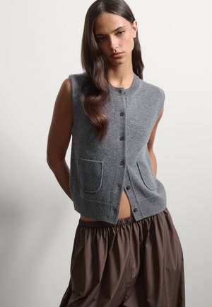 Jeune femme aux longs cheveux foncés porte un gilet en tricot gris sans manches avec boutons et un pantalon marron ample sur un fond clair uni.