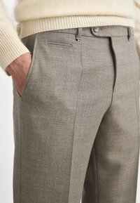 Beige textúrájú nadrág, testre szabott fazonnal, elülső zsebbel, gombos záródással és finom mintázattal.