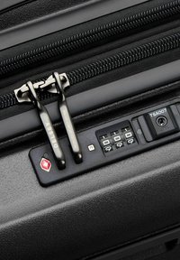 Valigia rigida nera con chiusura a cerniera, dotata di doppi tirazip in metallo nero, lucchetto TSA e meccanismo di chiusura a combinazione.