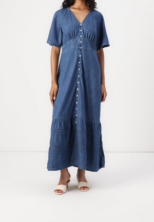 Kobieta w niebieskiej, długiej sukience jeansowej z krótkimi rękawami i guzikami z przodu, zestawiona z beżowymi sandałami na obcasie z odkrytymi palcami.