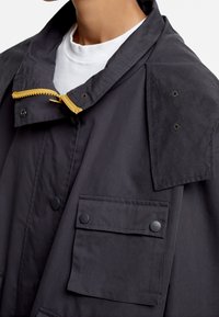 Veste imperméable noire en tissu lisse, dotée d'un col montant, d'une fermeture éclair jaune et de deux poches à rabat boutonnées.