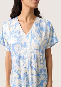 Ljusblå blus med blommönster, V-ringning, korta ärmar och en mjuk, slät textur. Har delikata vita blommönster.