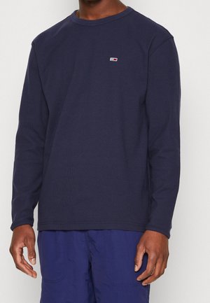 T-shirt à manches longues bleu marine en texture côtelée, avec un col rond et un petit logo cousu sur la poitrine gauche. Design simple et de couleur unie.