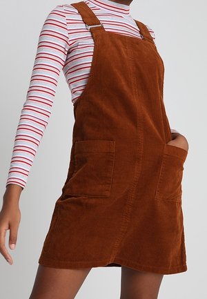 Persona que lleva un vestido pichi de pana marrón sobre una camisa blanca de manga larga con rayas horizontales rojas y azules.