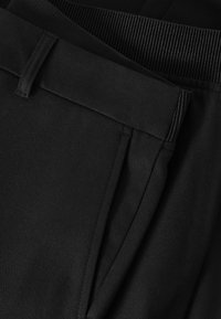 Gros plan sur un pantalon noir montrant la ceinture, les passants de ceinture et la poche latérale avec des coutures visibles et la texture du tissu.