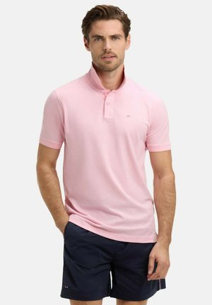 Homme portant un polo rose clair à manches courtes avec un col montant et un short bleu marine foncé, debout avec une main dans la poche.