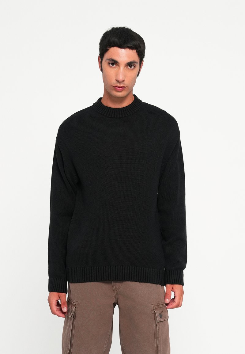 Pull en tricot noir avec un col rond côtelé, des manches longues et des poignets et un ourlet côtelés. Porté sur un pantalon cargo beige.