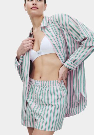 Vrouw die haar roze gestreepte pyjamashirt met groene strepen rechtzet, draagt bijpassende short en witte beha, staand tegen een effen achtergrond.