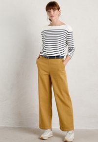 Top de rayas de manga larga en blanco y navy, combinado con pantalones de pierna ancha mostaza y un cinturón oscuro. Los zapatos son blancos con detalles en beige.