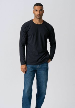 Mand står fremadrettet iført en sort langærmet skjorte og blå jeans med den ene hånd i lommen foran en ensfarvet grå baggrund.