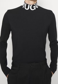 Schwarzes Langarmshirt mit hohem Kragen, das mit fettem weißen "HUGO"-Text gestaltet ist. Weicher Stoff und taillierte Passform. Kombiniert mit dunklen Hosen.