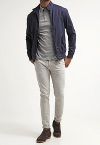 Veste légère bleu marine sur un polo gris, associée à un pantalon ajusté gris clair et des chaussures marron foncé. Design et textures simples.