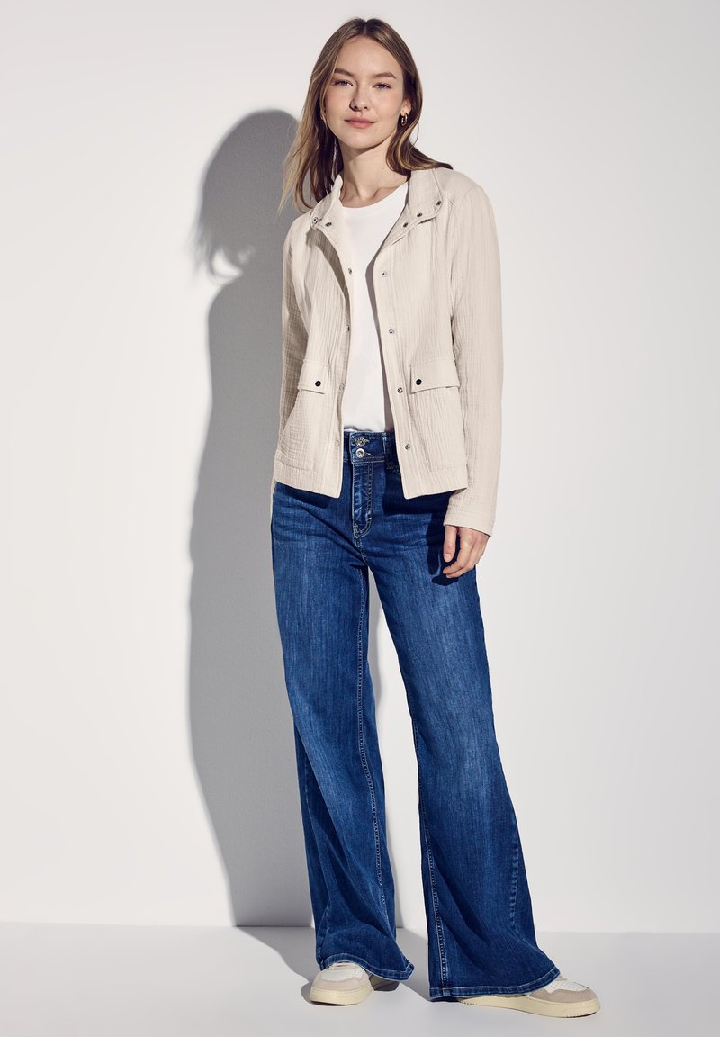Lichtbeige jas met een textuurafwerking, twee voorzakken, gecombineerd met blauwe flared jeans en beige schoenen. Eenvoudig, casual ontwerp en kleur.