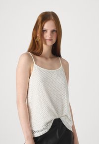 Hvit, heklet tanktopp med justerbare stropper, formet helning og et teksturert blomstermønster. Kombinert med sorte skinnbukser.