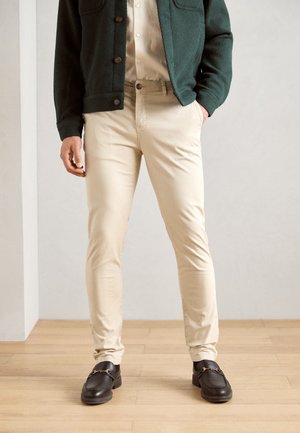 Light beige trousers paired with a dark green textured cardigan, featuring large brown buttons and black loafers with a gold accent.