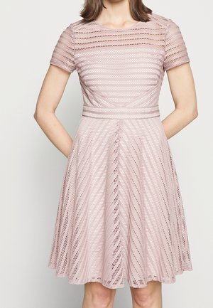 Donna che indossa un vestito rosa fino al ginocchio, a maniche corte, con motivo a pizzo a righe diagonali e vita aderente, in piedi con le mani dietro la schiena.