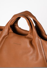 Braune Leder-Tote mit strukturiertem Oberflächen, gefaltetem Design und doppelten Griffen. Besitzt ein dezentes, geprägtes Logo auf der Vorderseite.