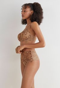 Completo di lingerie in pizzo nei toni nudi, caratterizzato da motivi floreali, spalline regolabili, bordi scallop e un design a vita alta.