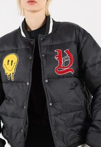 Black puffer jacket with striped collar, featuring a yellow smiley face and red embroidered letter "U" on the front. Textured fabric, button closure.