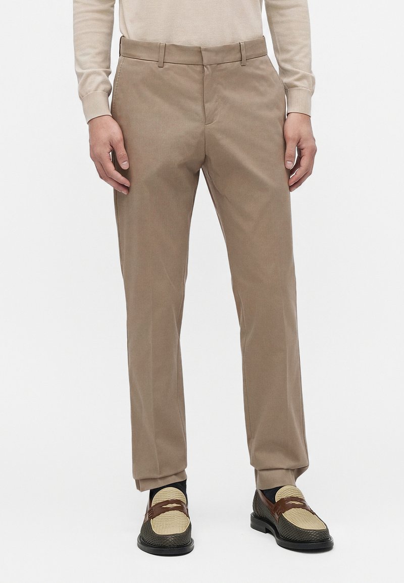 Homme portant un pantalon beige droit, un haut crème à manches longues, des mocassins en cuir tissé noir et beige, debout sur un fond blanc.