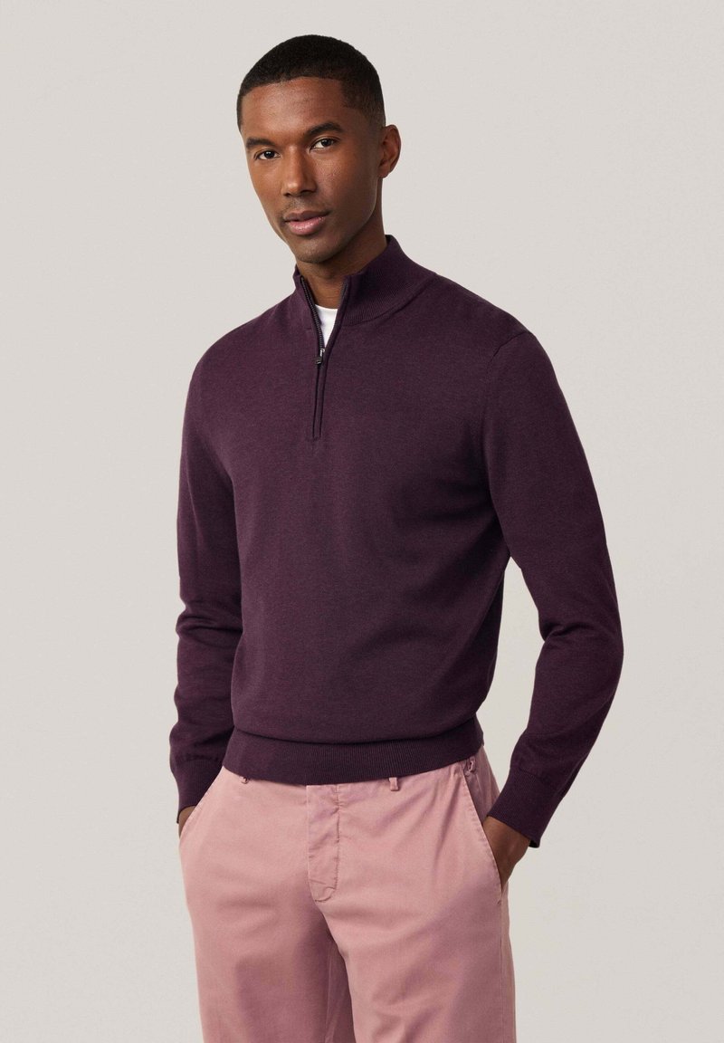 Pull-over à col zippé violet foncé avec des manches longues et un ourlet côtelé ; porté avec un chino rose clair, comportant des poches avant.