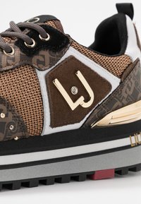 Zapatilla deportiva con parte superior de malla marrón, acentos de ante y logo metálico dorado. Entresuela gruesa gris con suela exterior de goma negra y detalle en rojo.