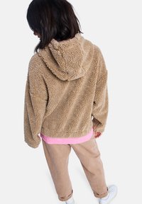 Beige fleece hoodie met een textuur van een pluizige oppervlakte, voorzien van een capuchon en een roze shirt daaronder. Beigekleurige broek en witte schoenen zijn zichtbaar.