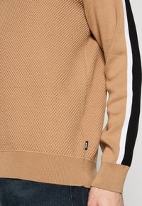 Pull en maille couleur chameau avec devant texturé, rayures noires et blanches sur les manches, porté avec un jean foncé, avec une petite étiquette "BOSS" sur l'ourlet.