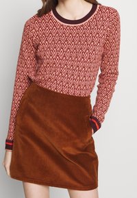 Langärmliger roter und cremefarbener Pullover mit einem geometrischen Muster, kombiniert mit einem braunen Baumwollröckchen aus Cord mit seitlichen Taschen.