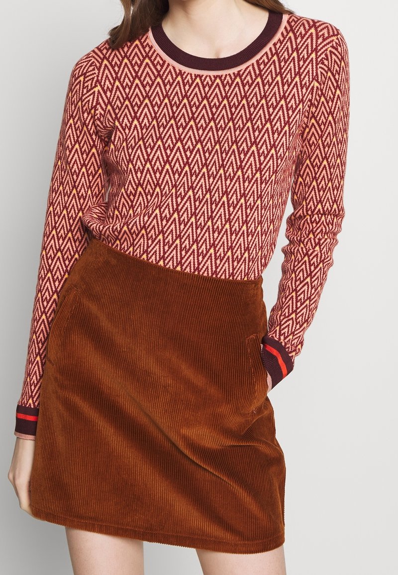 Langärmliger roter und cremefarbener Pullover mit einem geometrischen Muster, kombiniert mit einem braunen Baumwollröckchen aus Cord mit seitlichen Taschen.