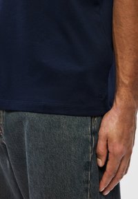 Meia-cintura de uma pessoa vestindo uma camisa azul marinho e jeans escuros, com a mão visível descansando ao longo da coxa.
