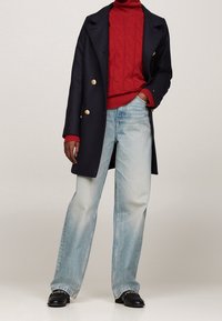 Marineblauer doppelt knöpfbarer Mantel mit goldenen Knöpfen, rotem Kabelstrick-Pullover mit Rollkragen, hellblauen geraden Jeans und schwarzen Schuhen mit dekorativen Akzenten.