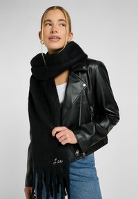 Écharpe en laine noire avec des extrémités frangées, comportant un petit logo brodé. Portée avec une veste en cuir noire et un haut blanc.