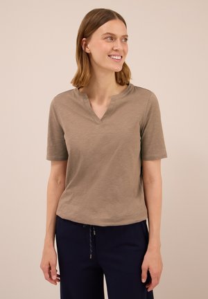 Donna sorridente, che indossa una maglietta beige a maniche corte con scollatura a V e pantaloni blu scuro con coulisse, su uno sfondo semplice.