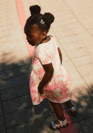 Ung jente med to hårboller iført en blomstermønstret kjole og sølvfargede sandaler som står på belegningsstein nær en rød linje i sterkt sollys.