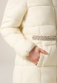 Krämfärgad quiltad vadderad jacka med midjebälte prytt med en dekorativ kristalldetalj och sidoficka. Mjuk textur och slät yta.