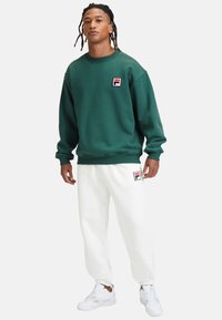 Felpa verde con polsini e orlo a costine, con un piccolo logo rosso e bianco, abbinata a pantaloni della tuta bianchi e sneaker bianche.