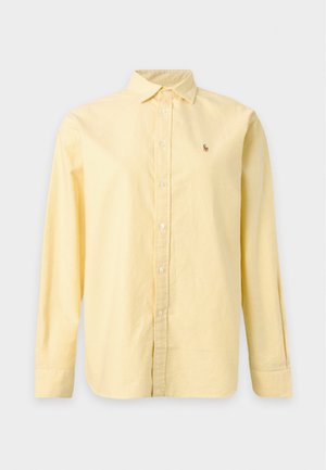 Femme debout, main dans la poche, portant une chemise jaune ample boutonnée, un jean bleu foncé et des sandales plates noires sur fond blanc.