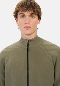 Olivengrøn zip-up sweater med høj krave og tekstureret strikstof. Har sort lynlås og raglanærmer for øget komfort.
